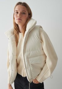 Dik, crèmekleurig vest met een nep-shearling kraag, ritssluiting en zijzakken, gelaagd over een licht gebreid trui.