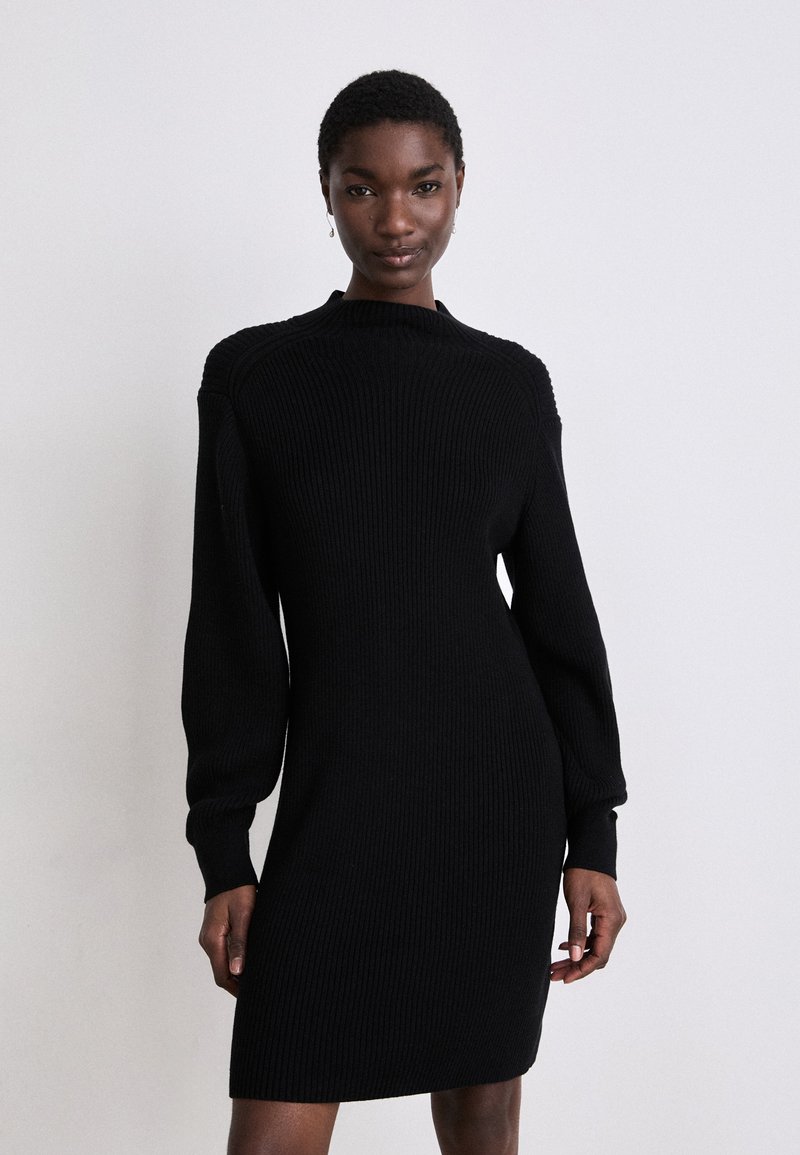 Vestido preto de malha canelada com mangas longas, gola alta e silhueta ajustada. O material tem um acabamento texturizado. Design simples e elegante.