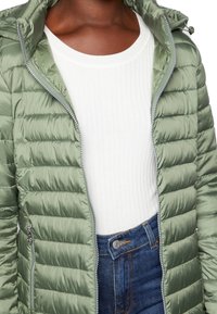 Lys grøn pufferjakke med lynlås og horisontalt quiltet mønster, båret over en hvid ribbestrikket sweater og blå jeans.