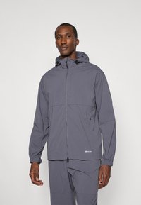 Giacca impermeabile grigia con cappuccio, zip frontale, polsini elastici e dettagli con coulisse. Il tessuto appare liscio e leggero.