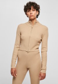 Ribbad beige zip-up tröja med hög krage och långa ärmar, med en figurnära design och textilstruktur.