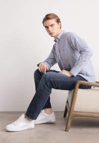 Camisa de botones a rayas azul y blanca, jeans de mezclilla oscuros y zapatillas blancas, sentado en una silla de color claro con patas de madera.