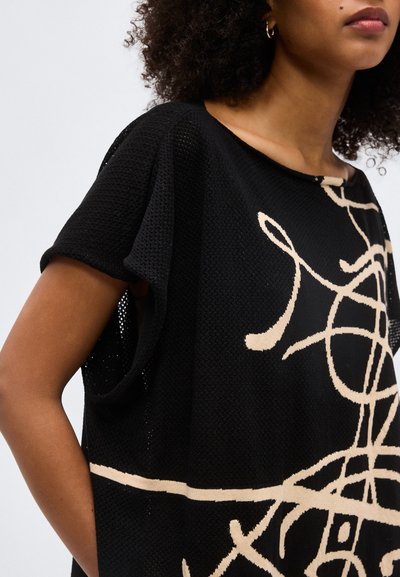 Γυναίκα που φοράει μαύρη πλεκτή μπλούζα με μπεζ αφηρημένο γραμμικό μοτίβο και κοντά μανίκια, εμφανίζεται από τον ώμο ως τη μέση σε απλό φόντο.