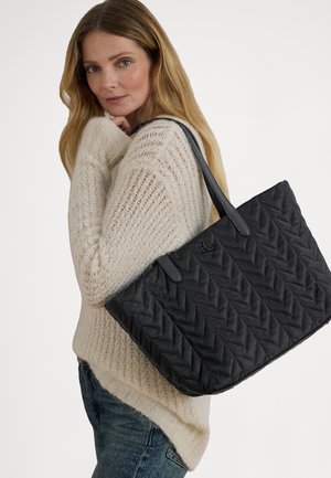 Sort quiltet shopper-taske med geometrisk mønster, der har to håndtag og en tekstureret overflade. Kombineret med en lys strikket sweater.