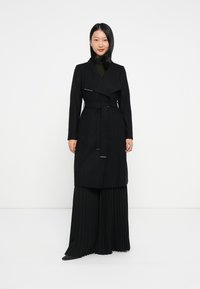 Casaco trench preto com um design envolvente, detalhe de cinto e um colarinho. O material parece texturizado. Usado sobre calças pregueadas pretas.