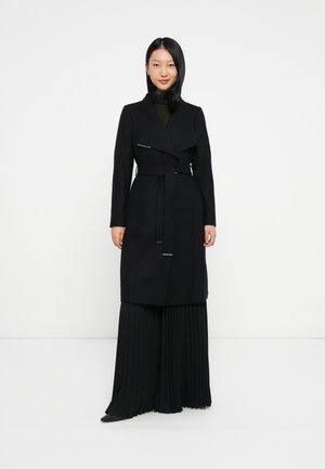 Abrigo negro tipo trench con un diseño envolvente, detalle de cinturón y un cuello. El material parece tener textura. Lleva puesto sobre unos pantalones negros de pinzas.