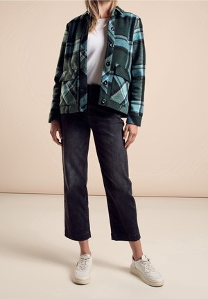 Groene geruite jas met blauwe accenten, twee voorzakken, zwarte rechte spijkerbroek en witte sneakers. Soepele textuur met knoopsluiting.