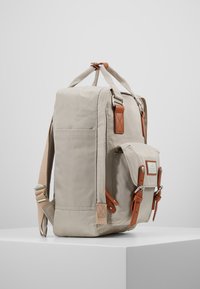 Sac à dos en toile beige avec accents en cuir, poche latérale, bretelles ajustables et quincaillerie en métal. Forme carrée avec coins renforcés.