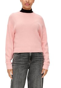 Pull en maille rose à col montant et épaules tombantes, avec des manches longues et un design court. Associé à un jean noir.