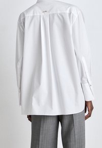 Camicia bianca a maniche lunghe con vestibilità rilassata, piega sul retro e dettaglio del logo dorato sul collo. Presenta polsini con bottone e una texture in cotone liscia.