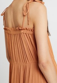 Kvinna som bär en ärmlös klänning i rostorange med samlad tyg och knutna axelband, visad från bak- och sidorna.