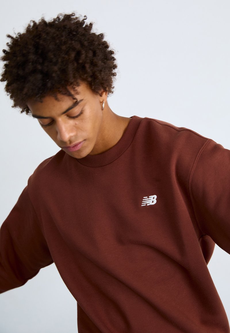 Brauner Sweatshirt mit rundem Ausschnitt, gerippten Bündchen und Saum. Verfügt über ein kleines weißes New Balance-Logo auf der Brust. Glatte Textur.