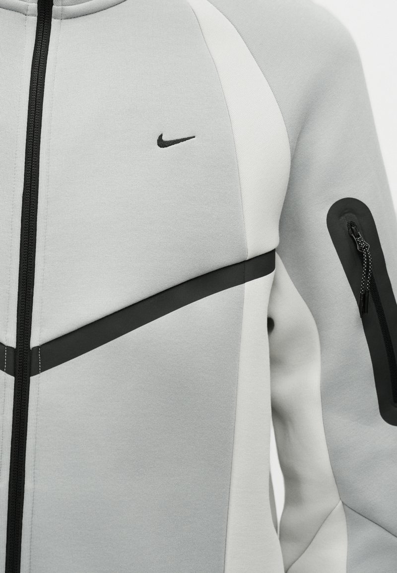 Hellgrauer Kapuzenpullover mit Reißverschluss und schwarzem diagonalen Streifen über der Brust, kleinem schwarzem Nike-Swoosh auf der linken Brust und Reißverschlusstasche am linken Ärmel.