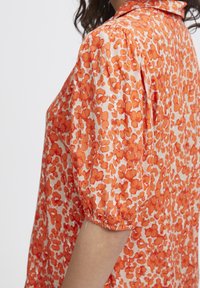 Short-sleeved blouse in orange floral print on a light background. Features gathered sleeves and a relaxed fit. Smooth texture, lightweight fabric.
