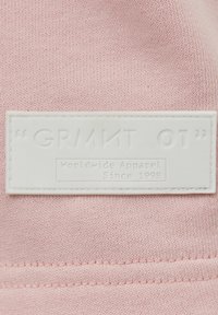 Étiquette de vêtement rectangulaire blanche sur tissu rose avec le texte en relief « GRMNT OT » et un texte plus petit « Vêtements du monde depuis 1998. »