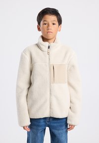 Cream-colored fleece jacket with a high collar, front zipper, and a beige chest pocket. Soft texture and loose fit. Worn over denim jeans.