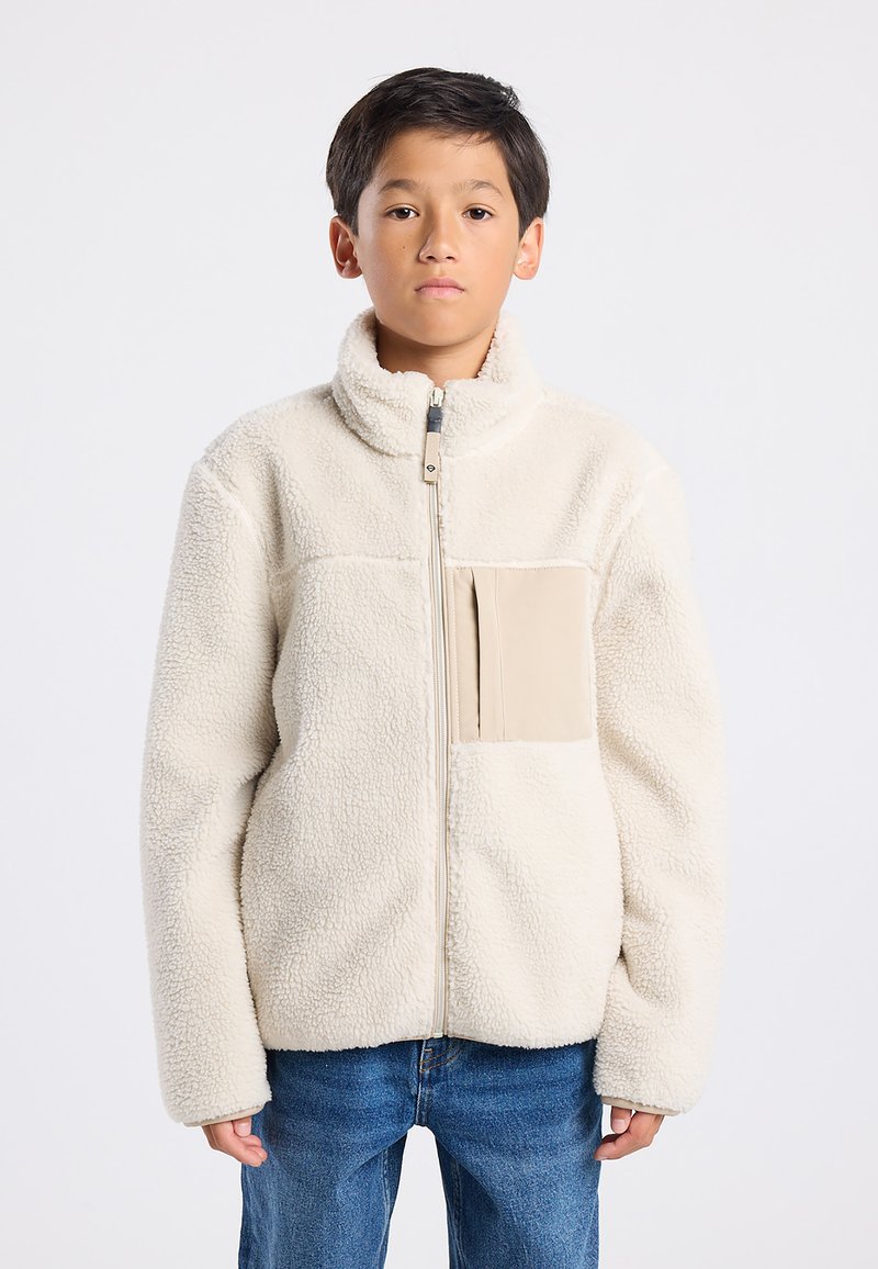 Cream-colored fleece jacket with a high collar, front zipper, and a beige chest pocket. Soft texture and loose fit. Worn over denim jeans.