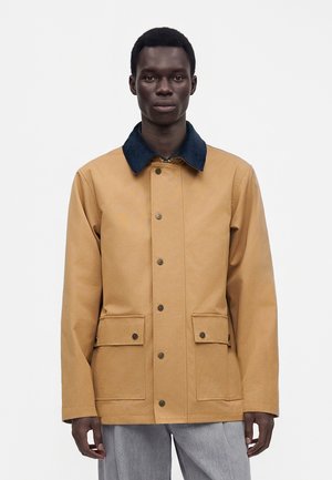 Homme face à l’avant portant une veste beige à col noir, fermetures à boutons et poches à rabat sur le devant, associée à un pantalon gris clair.