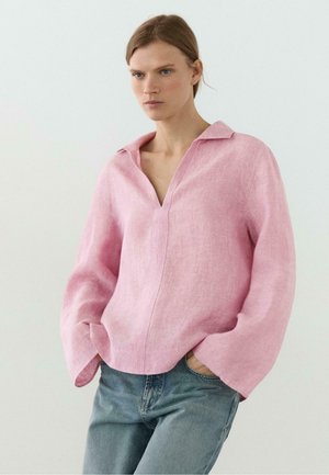 Kobieta o blond włosach, ubrana w luźną różową lnianą bluzkę z szerokimi rękawami i niebieskie dżinsy, stoi z rękami w kieszeniach.