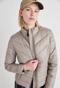 Lett beigepufferjakke med chevron-søm, høy krage og glidelås foran. Myk, glatt tekstur; tettsittende design.