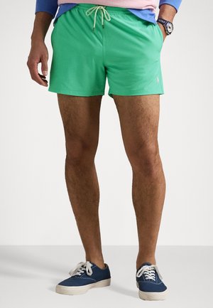 Bañador verde hecho de tela de secado rápido, con una cinturilla elástica y cordón. Combinado con zapatillas de lona azul oscuro.
