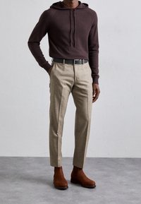 Man med mörkbrun huvtröja, skräddarsydda beiga byxor med ett svart bälte och bruna mocka Chelsea-boots, stående mot en enfärgad bakgrund.