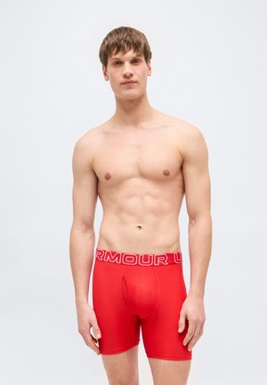 Νέος άνδρας όρθιος, χωρίς πουκάμισο, φορώντας κόκκινα εσώρουχα Under Armour boxer, ουδέτερη έκφραση, απλό φωτεινό φόντο.