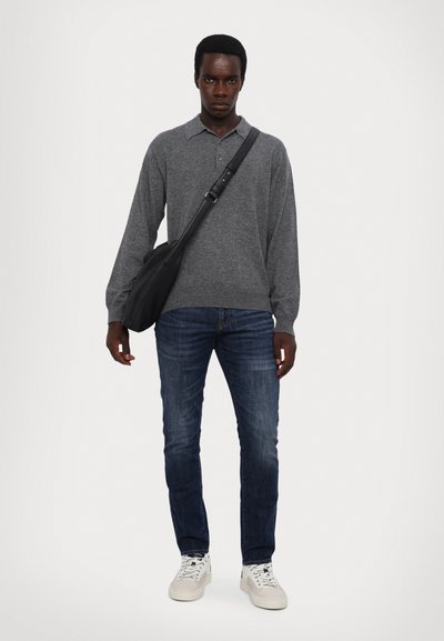 Graues Langarm-Polo-Shirt, blaue Jeans und weiße Sneakers. Schwarze Umhängetasche. Modell steht vor einem weißen Hintergrund.