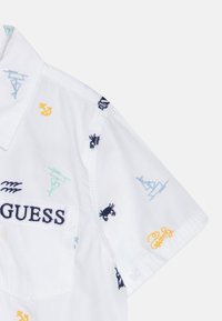 Camicetta bianca a maniche corte con vari motivi nautici ricamati, tra cui granchi e ancore, e il logo "GUESS" in blu navy.