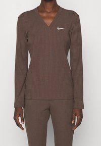 Haut à manches longues marron côtelé avec un décolleté en V et un petit logo Nike blanc sur la poitrine. Tissu doux avec une silhouette ajustée.