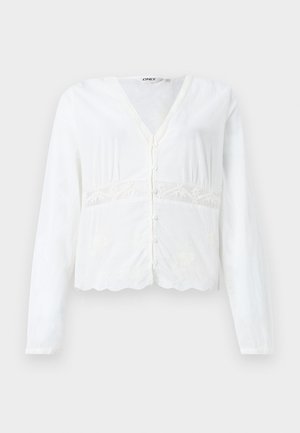 Λευκό πουκάμισο με κουμπιά και μακριά μανίκια, V-λαιμόκοψη, δαντελένιες λεπτομέρειες γύρω από τη μέση και κυματιστό τελείωμα.