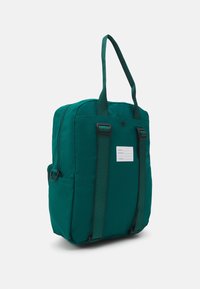 Sac à dos en tissu vert avec deux poignées supérieures, des sangles contrastées et une étiquette pour les informations personnelles. Il présente une forme structurée et une texture lisse.