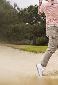 Golfista w różowej koszuli i beżowych spodniach wykonuje zamach kijem z piaskowej pułapki na zielonym polu golfowym z drzewami w tle.