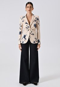 Crème blazer met bloemenprint en blauwe accenten, om de taille gebonden met een riem. Gecombineerd met hooggetailleerde donkere denimbroeken en glanzende zwarte hakken.