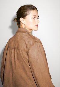 Brun ruskindjakke med krave, synlige sømme og afslappet pasform, vist fra siden med en minimalistisk baggrund.