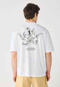 Hvit bomull T-skjorte med en svart illustrasjon av en fugl og blomster på baksiden, med trykt tekst under designet.