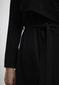 Robe portefeuille noire à manches longues, tissu texturé, taille cintrée avec une ceinture assortie et poches latérales. Lignes lisses et épurées sur toute la longueur.