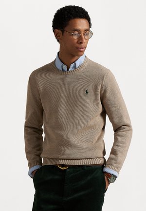 Beige knit sweater with ribbed crew neck and cuffs, featuring a small green logo. Worn over a light blue collared shirt and dark green pants.