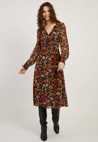 Vrouw met krullend haar, gekleed in een midi-jurk met lange mouwen met bloemen- en tijgerprint en zwarte kniehoge laarzen, glimlachend en staand voor een effen achtergrond.