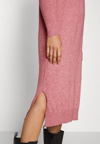 Robe en tricot à manches longues de couleur rose clair avec une fente sur le côté. Elle présente une texture douce et des poignets côtelés. Associée à des bottes noires à la cheville.