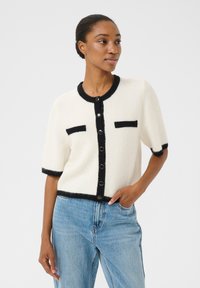 Short-sleeve cardigan in cream wool, featuring black trim and buttons, two black pocket accents, and a round neckline. Worn over denim jeans.