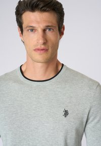 Maglione lavorato a maglia grigio chiaro con scollo rotondo, caratterizzato da un colletto a coste nero e un piccolo logo ricamato sul lato sinistro del petto.