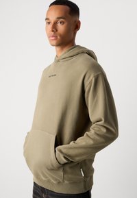 Khaki hoodie met een voorkantzak, ribgebreide manchetten en zoom. Kenmerkt zich door een subtiele logo op de borst. Zacht, gestructureerde stof.