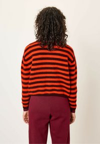 Pull rayé rouge et noir avec un tissu texturé, coupe décontractée et poignets côtelés, associé à un pantalon bordeaux. Vue de dos présentée.