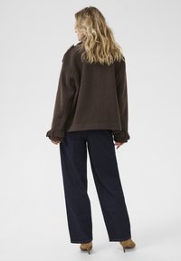 Femme aux cheveux blonds ondulés portant une veste marron, un pantalon large noir et des bottines à talons beiges, se tenant de dos sur un fond blanc.