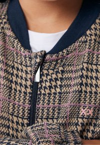 Pletená bomber bunda v béžovo-modrém houndstooth vzoru, se zipem vpředu a růžovým kontrastním prošíváním. Ribované límec a manžety.