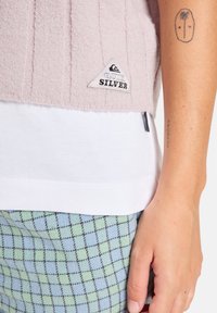 Haut en tricot flou rose clair avec une texture côtelée, superposé sur une chemise blanche. Associé à un pantalon à carreaux bleu et vert à motifs. Étiquette Quiksilver visible.