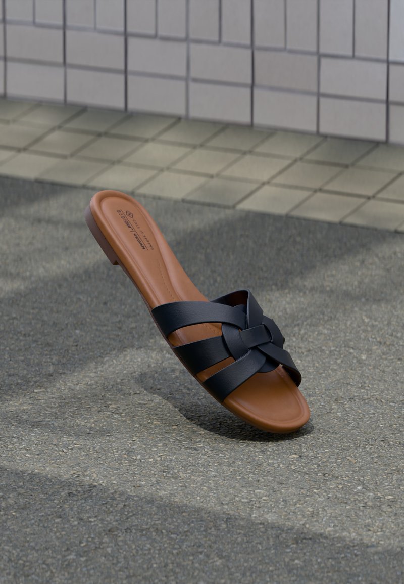 Καφέ επίπεδη σανδάλι με μαύρα πλεγμένα δερμάτινα λουριά τοποθετημένη όρθια σε άσφαλτο κοντά σε τοίχο με πλακάκια.