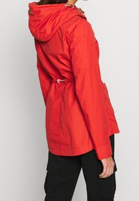 Veste rouge à capuche avec taille à cordon de serrage, poches latérales et texture lisse. Dotée d'une étiquette logo sur le haut du dos.