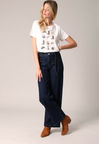 T-shirt graphique blanc avec diverses illustrations de bottes, associé à un jean taille haute bleu marine à jambes larges et des bottines en daim marron.